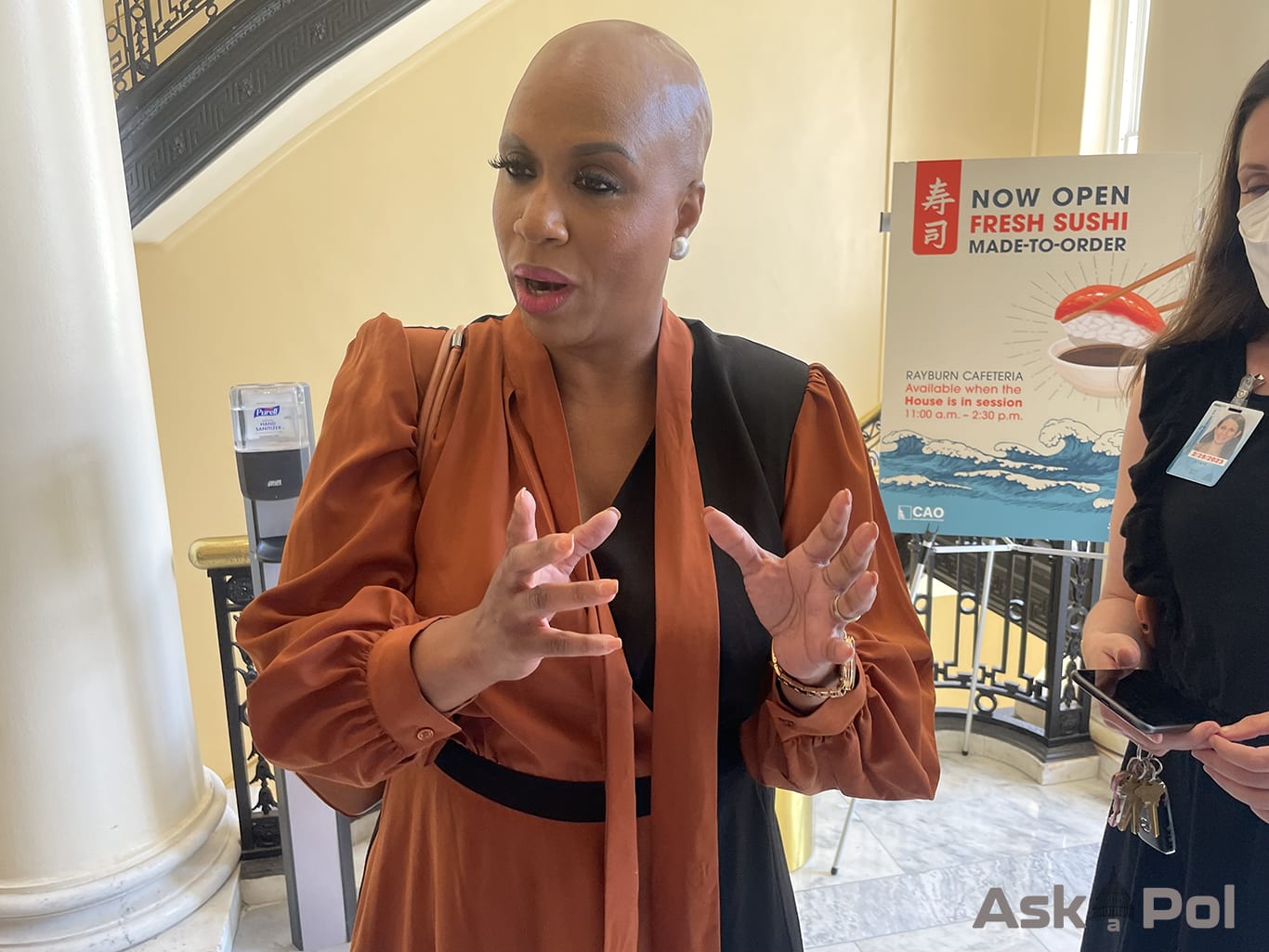 A bald African AMerican lady in an orange dress answers questions in a hallway near a staircase. Photo: Matt Laslo for www.askapol.com