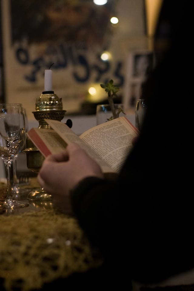 An image of a man's hands, holding a book open over a marble bar. Empty wine glasses and an unlit candle rest on the bartop in front of him.