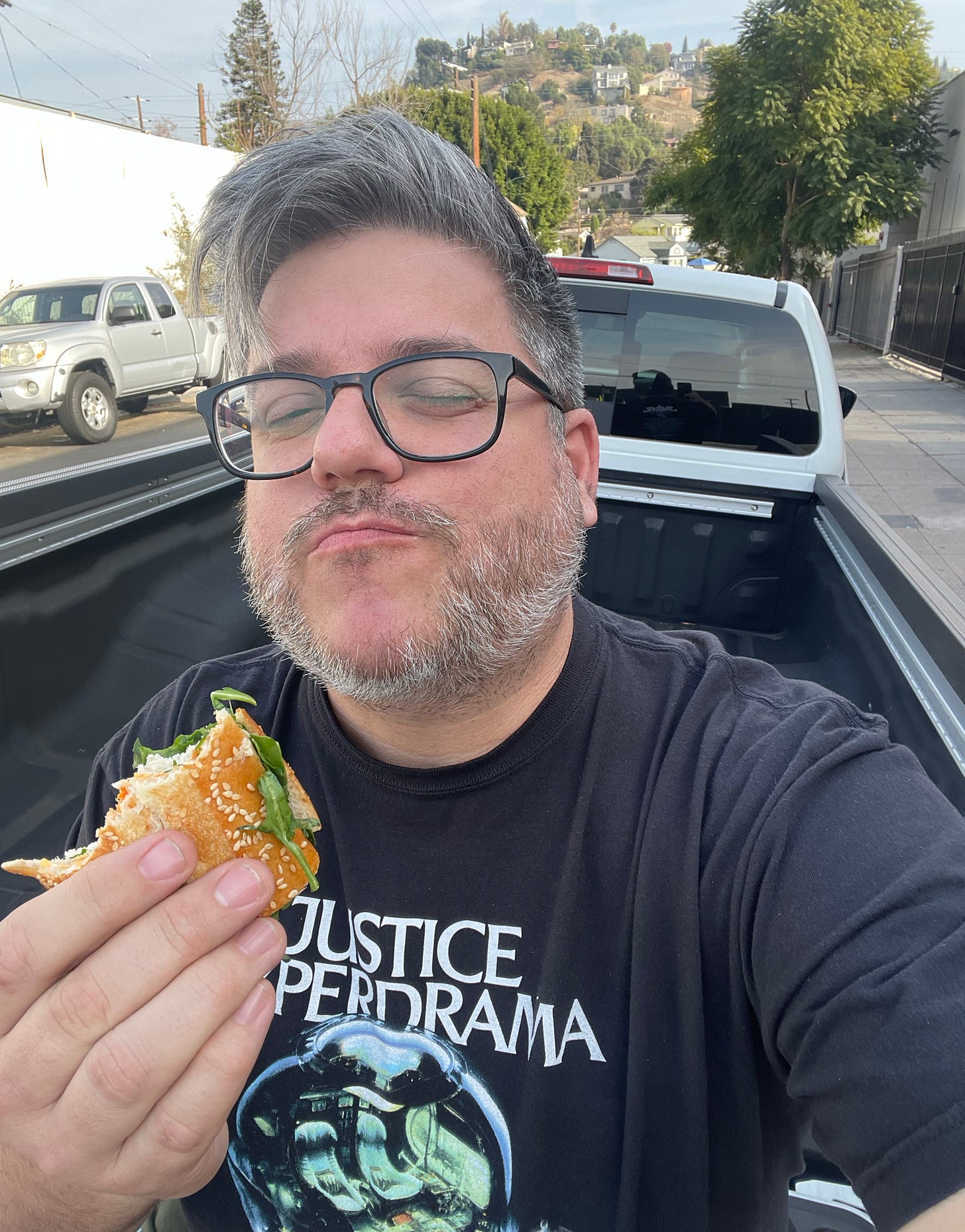Selfie of myself in the daytime, sitting in the back of a pickup truck with a sandwich in my hand. My eyes are closed and my face seems to express that I am having a great time.