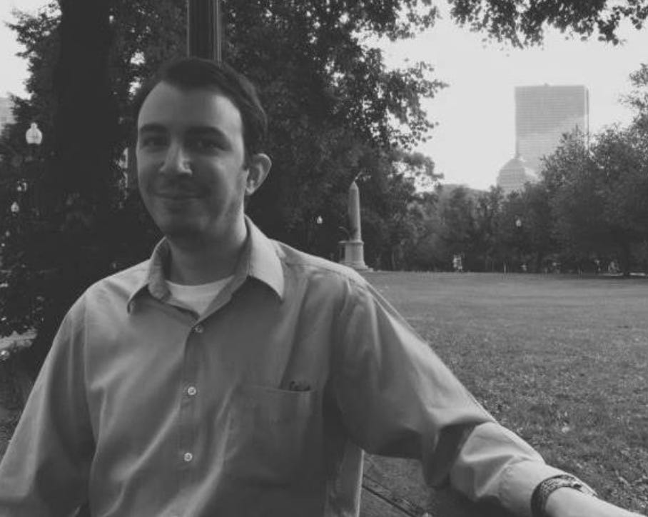 A white male sitting on a park bench. A patch of grass and the Boston skyline is behind his left shoulder; trees and a lightpole are behind his right shoulder. The photograph is in black and white.