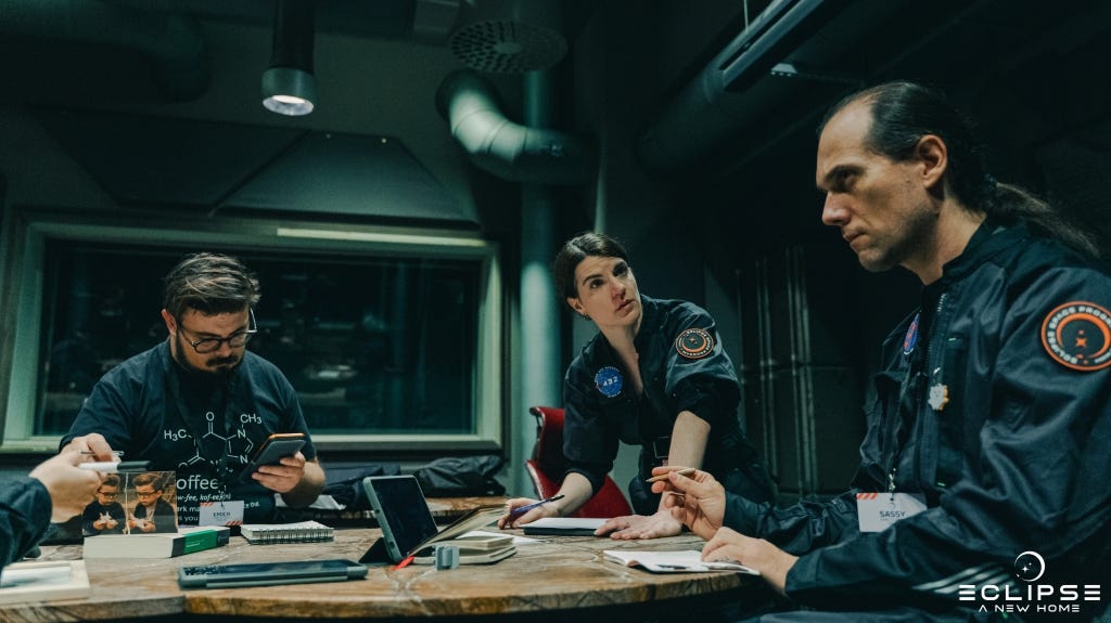 Three people sit around an office table, two of them in jumpsuits, consulting tablets
