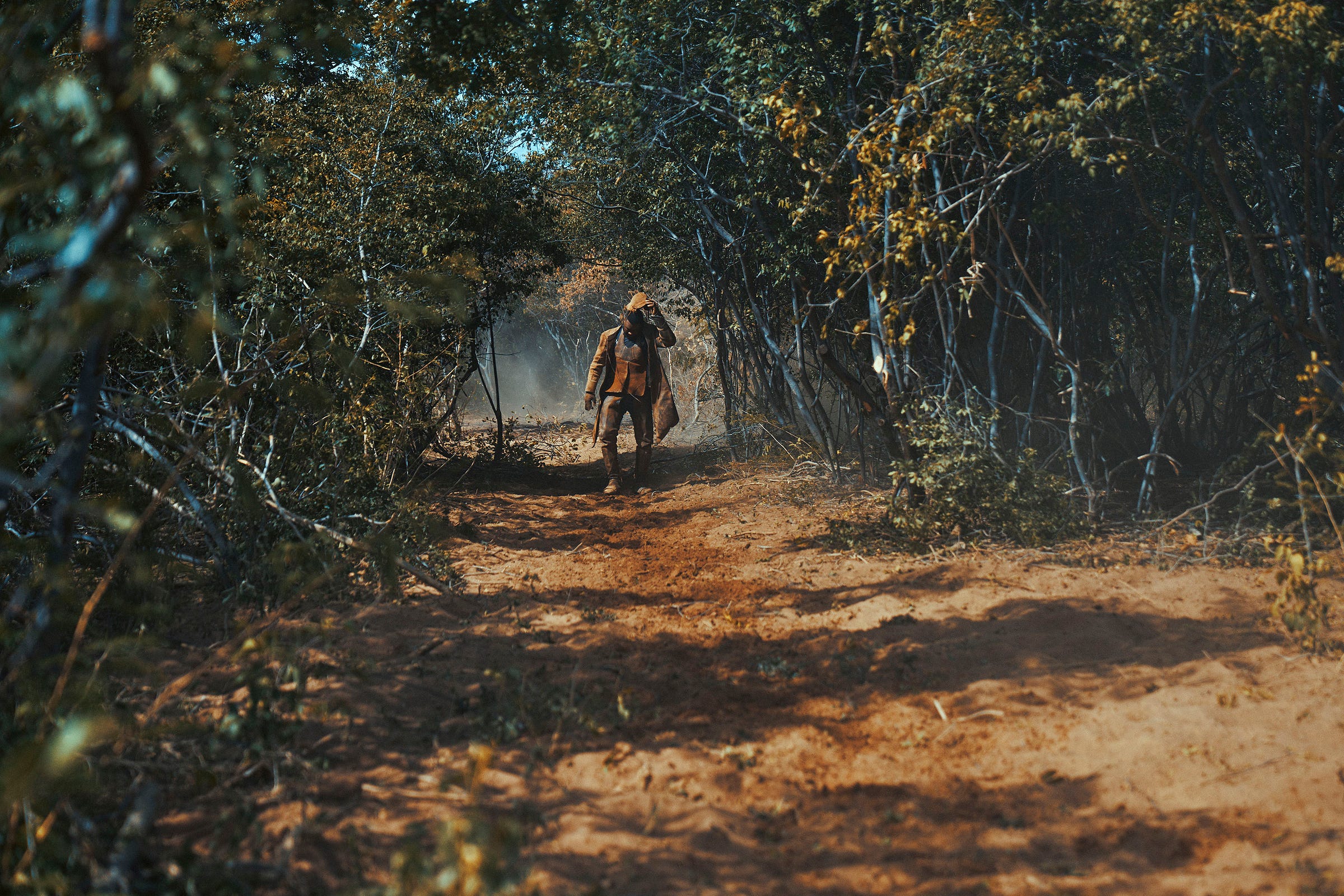 A person wearing outdoor gear and carrying a backpack walks down a sandy path surrounded by dense, green foliage and trees, with sunlight filtering through the branches. Dust rises along the trail.