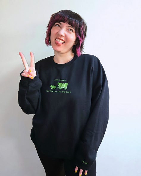 Modeling merch: A hot pink sweatshirt with white embroidered text on the front chest that says "gluten gives me the ick" and "GFGC" on the left sleeve near the wrist. A vintage wash denim baseball cap with an embroidered white pixelated chat bubble and black "GFGC" inside. A black sweatshirt with a green embroidered design on the front chest that says "GAME OVER, YOU ATE GLUTEN AND DIED" with a pixelated ox and wagon and "GFGC" on the left sleeve near the wrist.