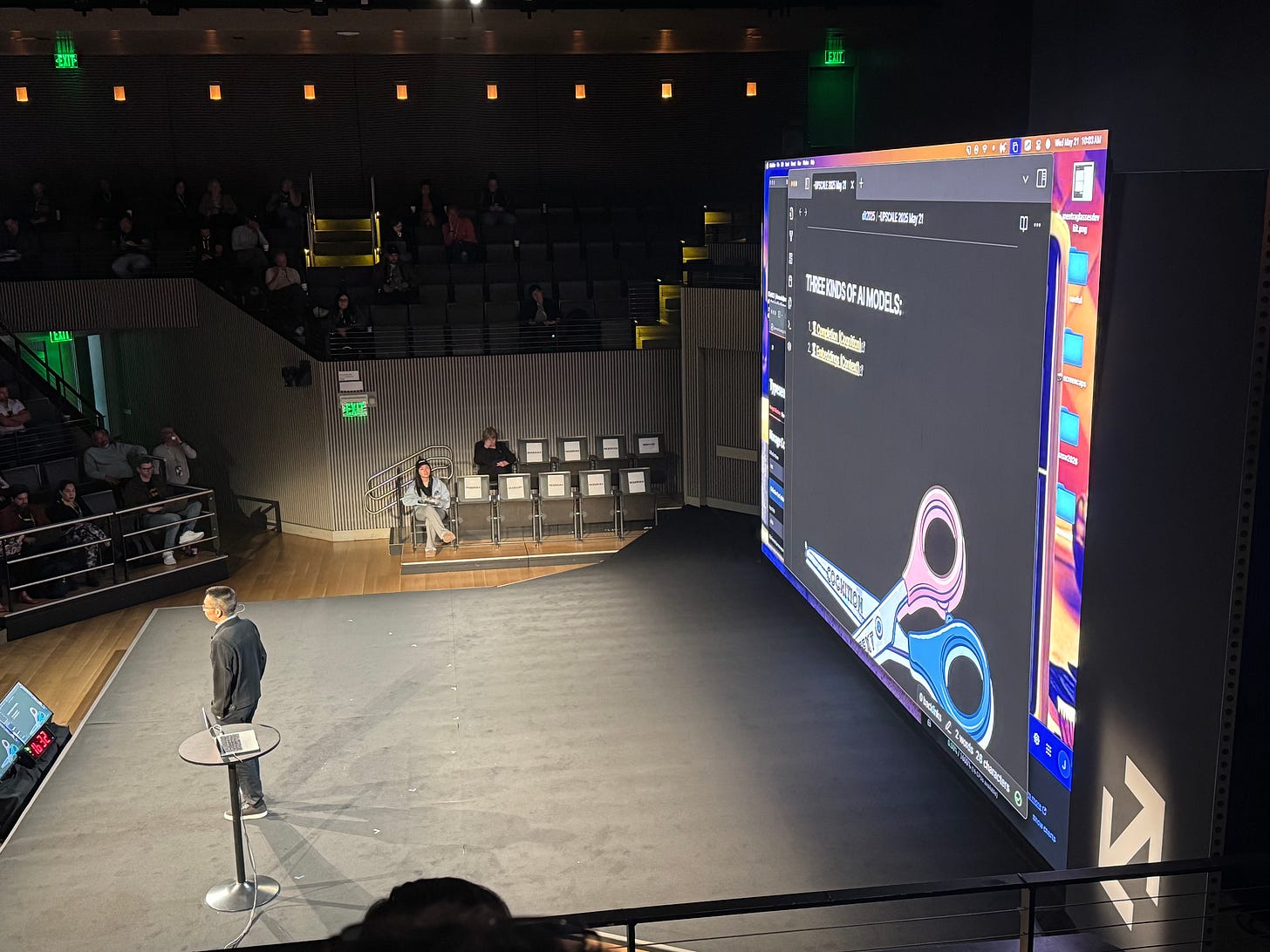 A speaker stands on stage at a conference next to a large screen displaying a slide titled "Three Kinds of AI Models." The slide features a visual metaphor of a pair of scissors, with one blade labeled "Cognition (Completion)" and the other "Context (Embeddings)." The audience is seated around the stage, and a laptop is placed on a small round table beside the speaker.
