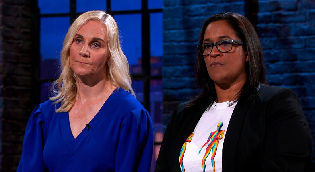 Two women - one with blonde hair and a blue top and one with brown hair and a blazer - looking into the distance with a window behind them Two women - one with blonde hair and a blue top and one with brown hair and a blazer - looking into the distance with a window behind them