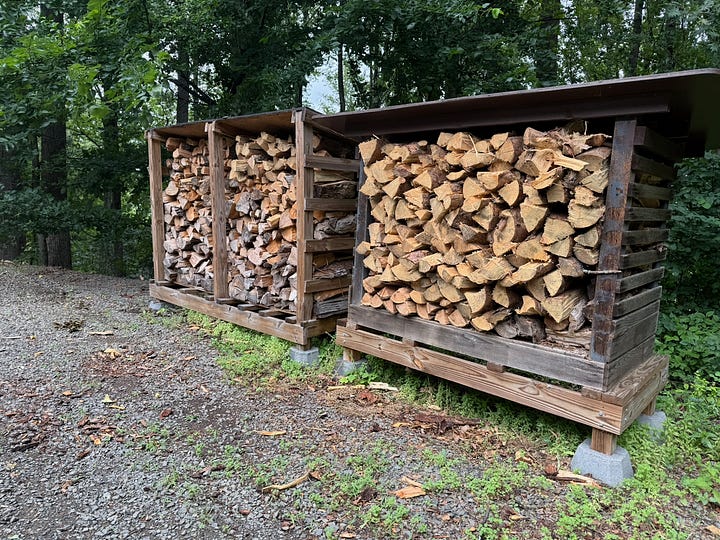 Two side-by-side photos documenting firewood processing and storage. The left image shows a work area beside a wooded tree line, with stacks of cut logs, a hydraulic log splitter, and freshly split firewood piled on the ground. The right image shows the finished result: three large, neatly stacked woodsheds filled with split firewood seasoning under metal roofs. Together, the images illustrate the transformation from raw logs to organized firewood storage.