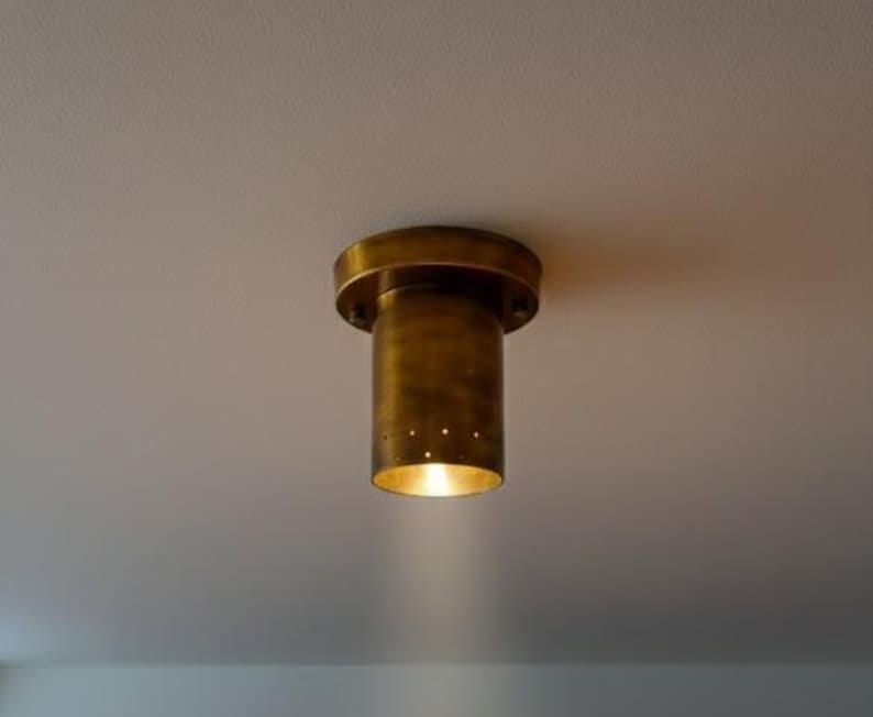 May include: A brass ceiling light fixture with a cylindrical shade and a circular base. The light is on, illuminating the space below. The fixture is mounted on a white ceiling, above a wooden floor. May include: A brass ceiling light fixture with a cylindrical shade and a circular base. The light is on, illuminating the space below. The fixture is mounted on a white ceiling, above a wooden floor.