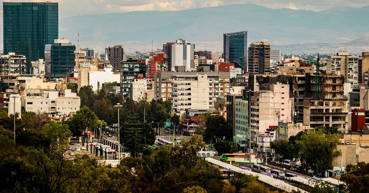 A wide view across central Mexico City: mid-rise apartment blocks and construction cranes, a Metrobús line running along a tree-lined avenue in the foreground, and the mountains ringing the valley behind.