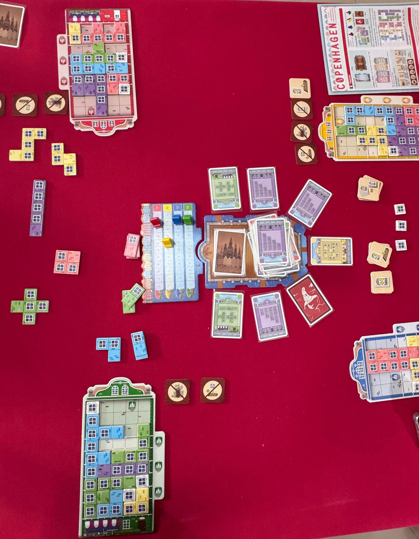 Uma mesa com toalha aveludada vermelha vista de cima. No meio há cartas e pequenos tabuleiros nas cores verde, vermelho, amarelo e azul. Por cima desses tabuleiros há pecinhas que lembram as do jogo Tetris, em várias cores e formatos, algumas com desenhos de janela em certos quadrados.