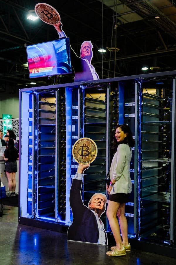 A woman poses with a cardboard cutout of President Trump holding a Bitcoin over his head. A woman poses with a cardboard cutout of President Trump holding a Bitcoin over his head.