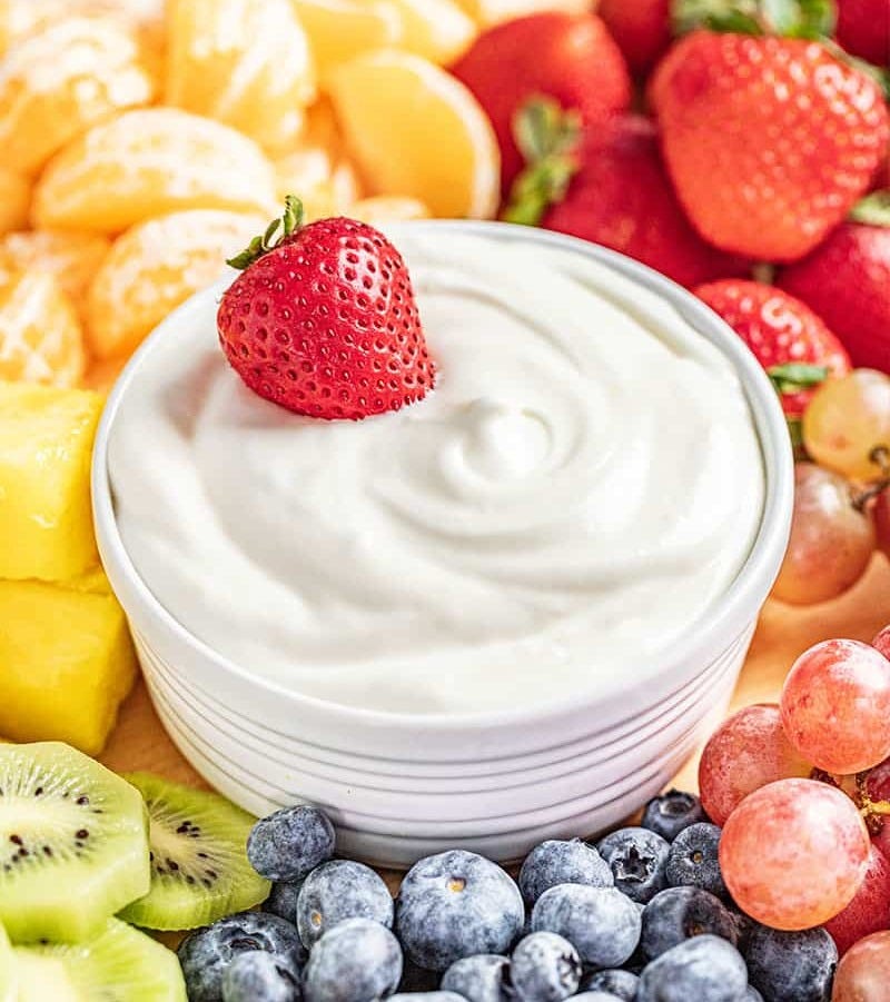 A close up view of a white bowl of fruit dip with a strawberry on top. The bowl sits in the middle of a fruit tray with a rainbow assortment of fruits.