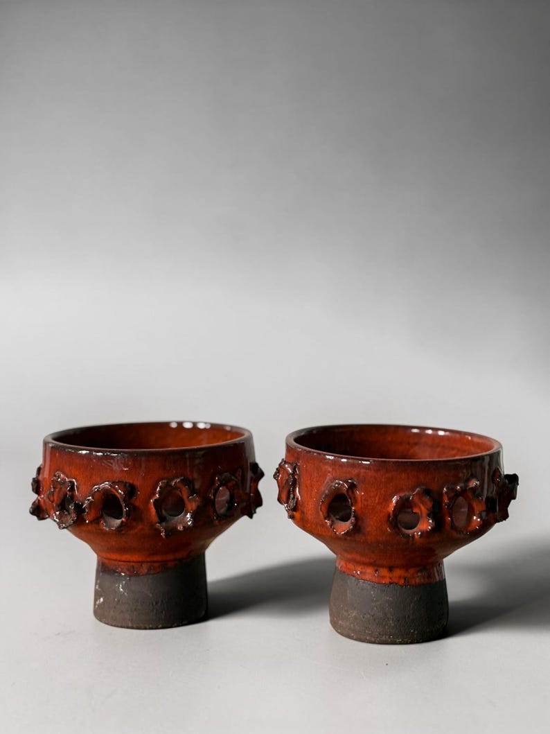 May include: Two matching ceramic bowls with a glossy, deep orange glaze on the upper sections and a matte, dark gray base. Each bowl features a series of circular, flower-like cutouts around the midsection, creating a textured, open-work design.