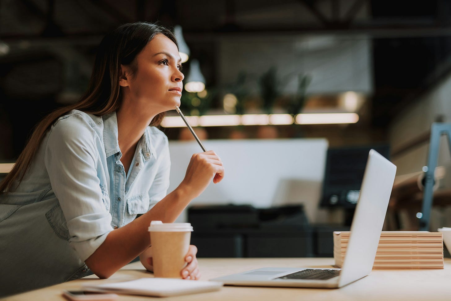 A thoughtful woman in a light blue shirt sits at a desk, holding a pencil beneath her chin while looking into the distance. An open laptop, notebook, and takeaway coffee sit in front of her in a warmly lit workspace. A thoughtful woman in a light blue shirt sits at a desk, holding a pencil beneath her chin while looking into the distance. An open laptop, notebook, and takeaway coffee sit in front of her in a warmly lit workspace.