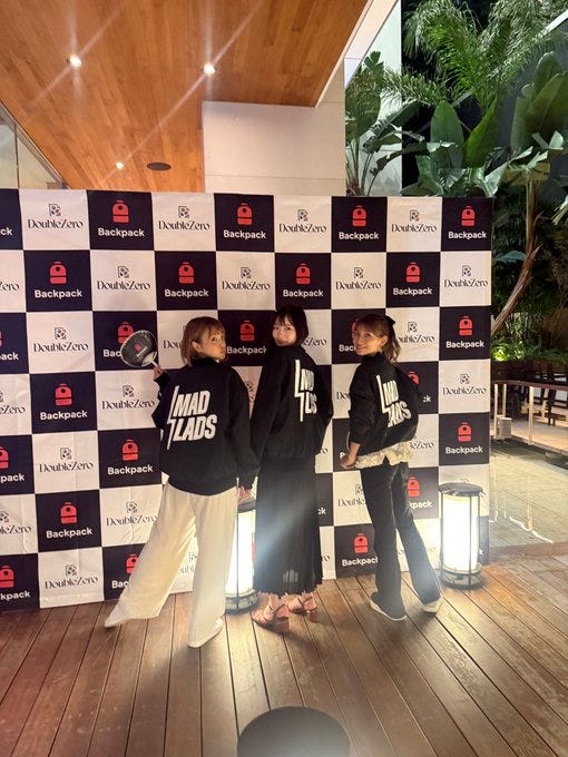 Three individuals standing on a wooden platform at night, wearing black and white clothing with "MadLads" text. They are positioned in front of a backdrop featuring Backpack and Double Zero logos in a checkered pattern. Tall plants and modern lanterns are visible in the background.