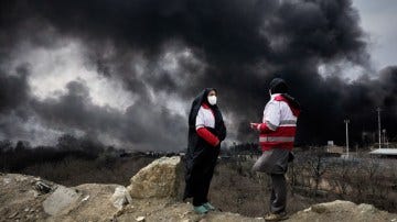 Two women from the Iranian Red Crescent Society stand on a dirt mound as a thick plume of smoke from an oil storage facility fire lingers in the sky over Tehran.