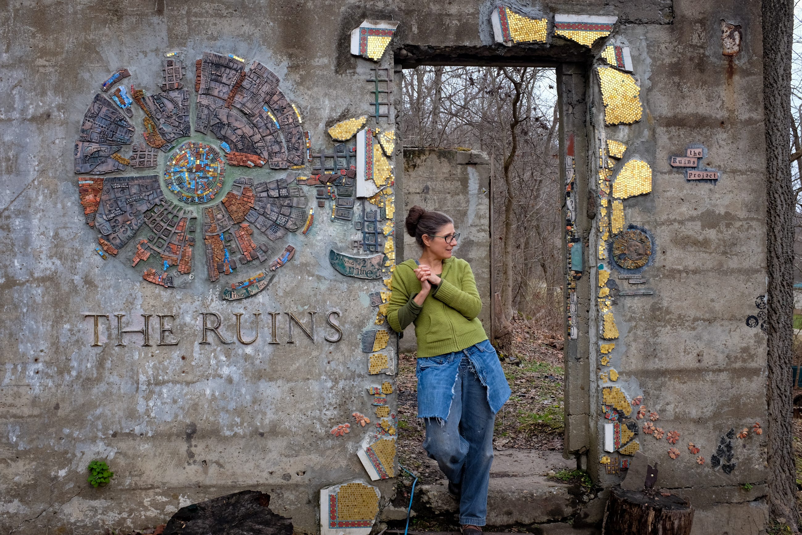 A white woman in jeans and a green hoodie with her hair in a bun stands with clasped hands in a doorway in a concrete wall. Mosaics surround the door. To the left of her there is a large round mosaic in gold and blues and browns with the words THE RUINS on the wall beneath it. A white woman in jeans and a green hoodie with her hair in a bun stands with clasped hands in a doorway in a concrete wall. Mosaics surround the door. To the left of her there is a large round mosaic in gold and blues and browns with the words THE RUINS on the wall beneath it.