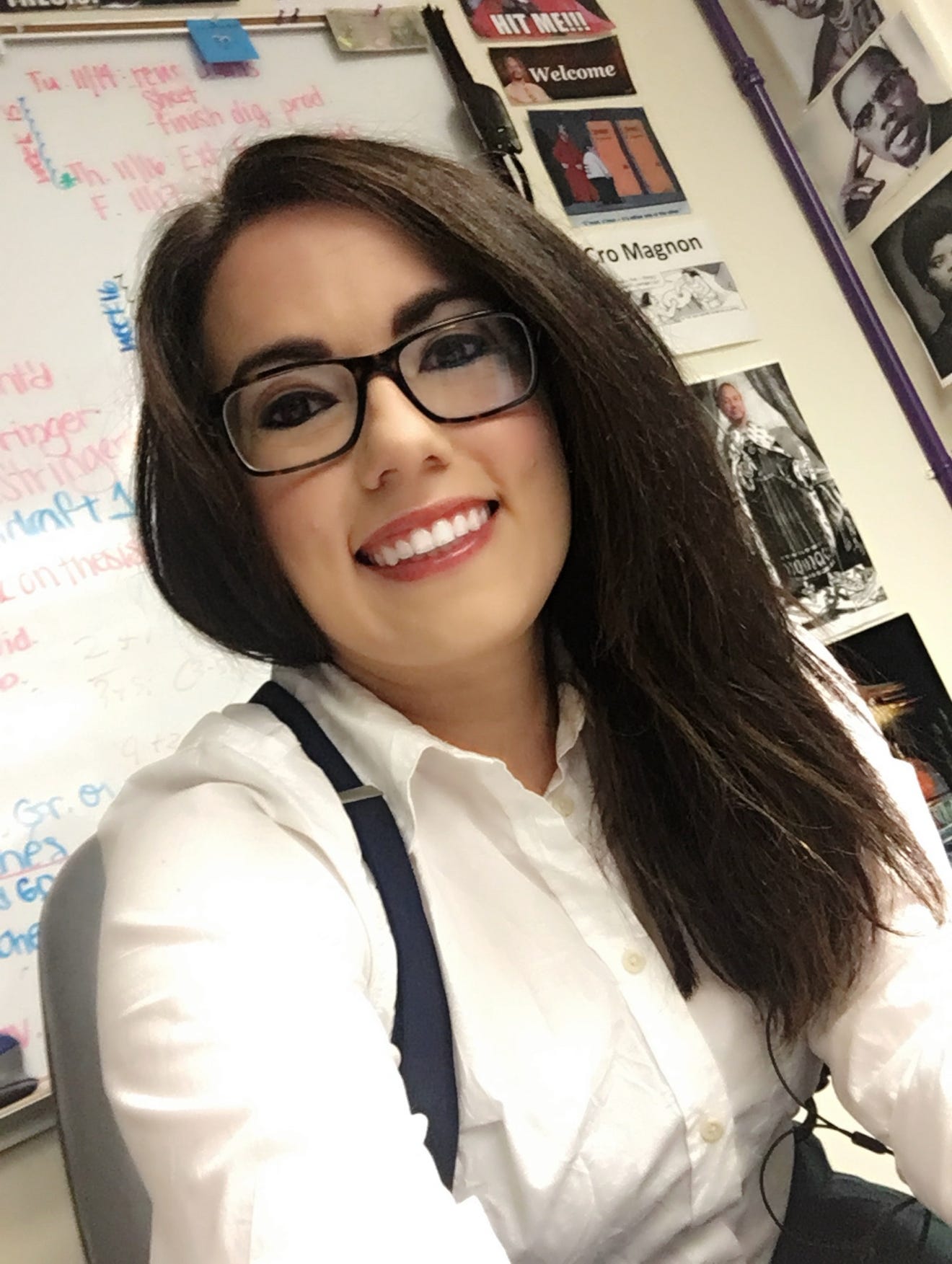 Andrea Gambino is smiling, sitting at a teacher desk. She has brown hair, brown eyes, and is wearing a white button down, glasses, and black suspenders!