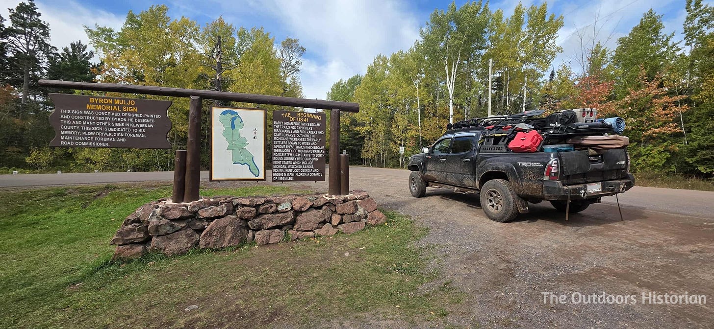 A black second generation Tacoma with roof racks and camping parked next to a wooden sign recognizing the beginning of US-41, including a map.