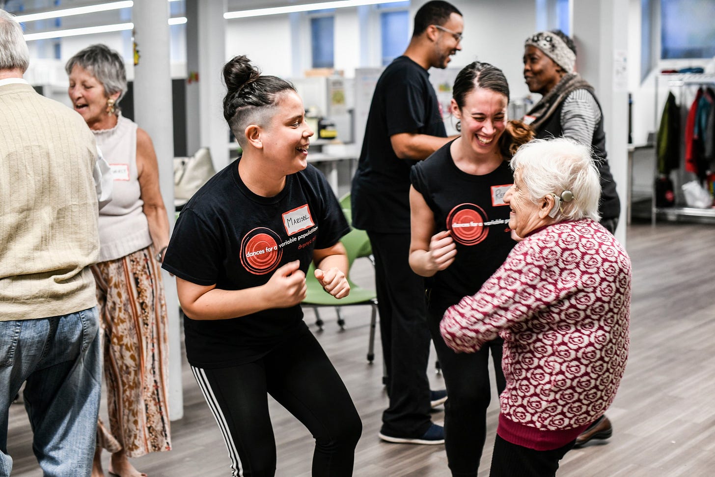 People socializing and dancing in an indoor space, smiling and laughing, including two young women in black shirts and an elderly woman in a patterned sweater.