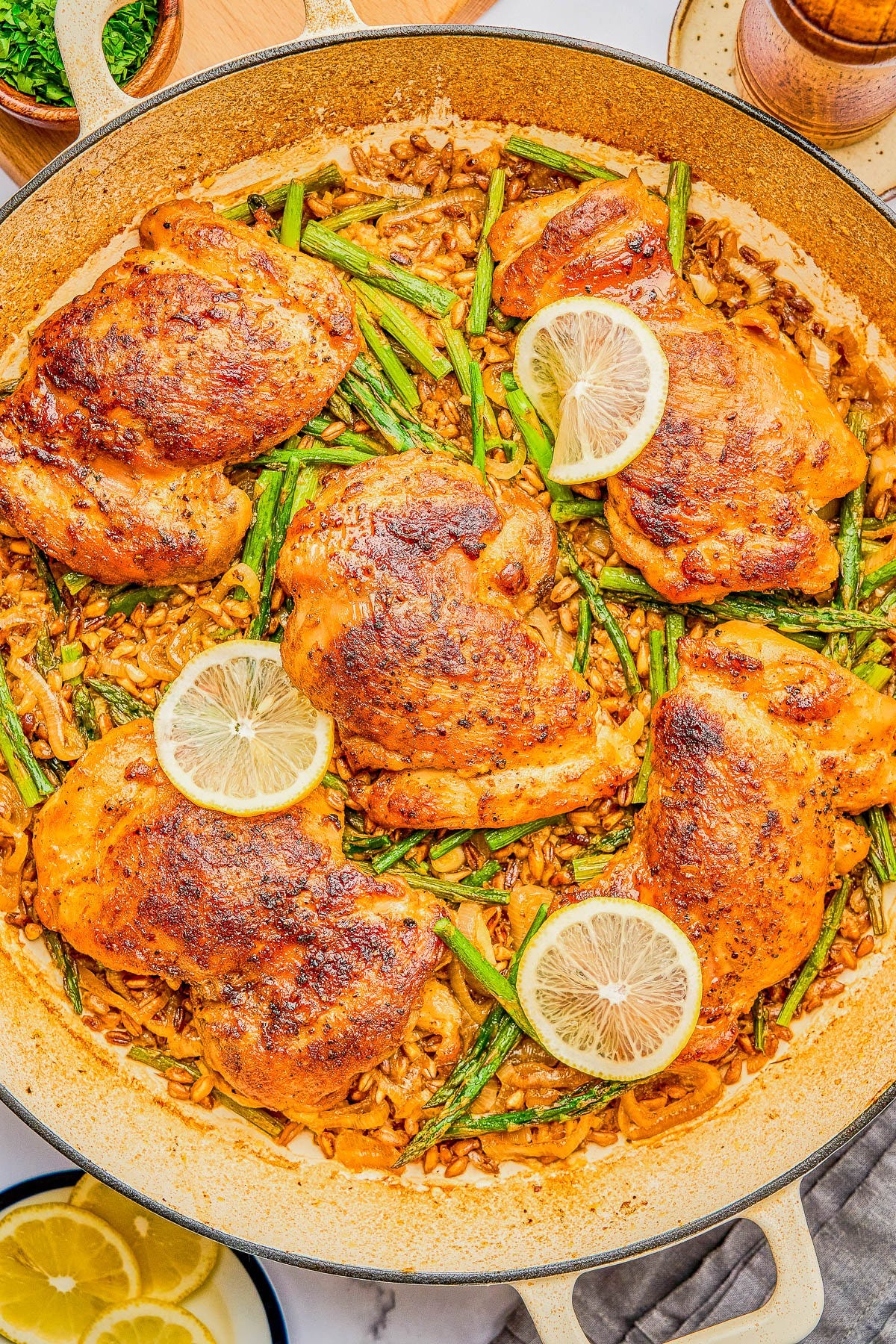 A large skillet with cooked chicken thighs, asparagus, lemon slices, and a rice dish, seasoned and arranged neatly.