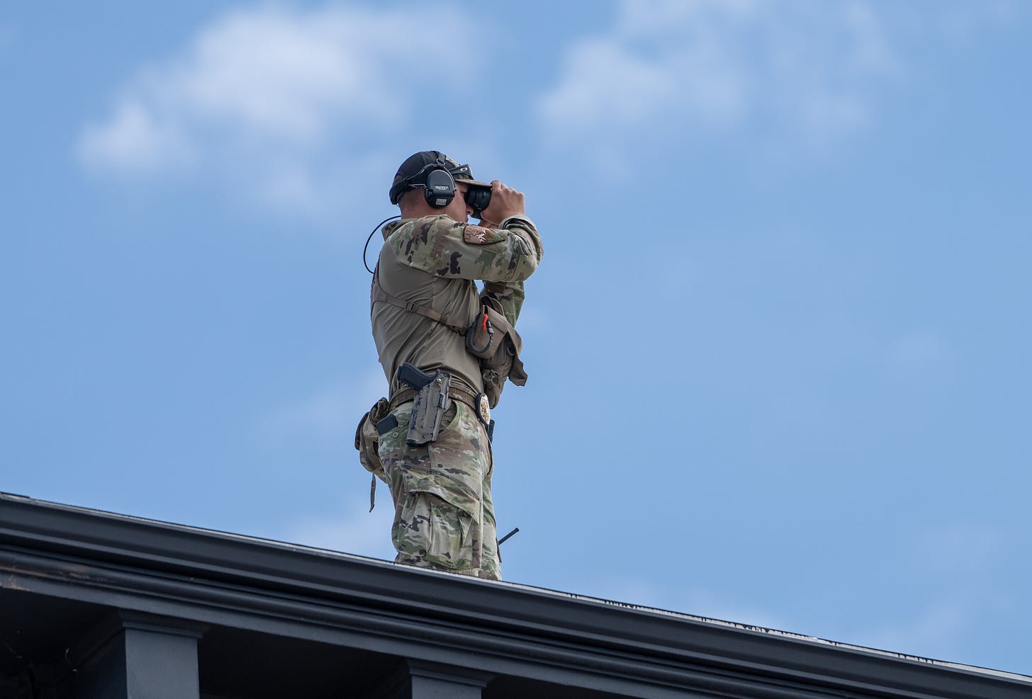 A man in camo with a gun on his belt peers through binoculars from his location on a downtown roof. Behind him the sky is a bright, clear blue. A man in camo with a gun on his belt peers through binoculars from his location on a downtown roof. Behind him the sky is a bright, clear blue.
