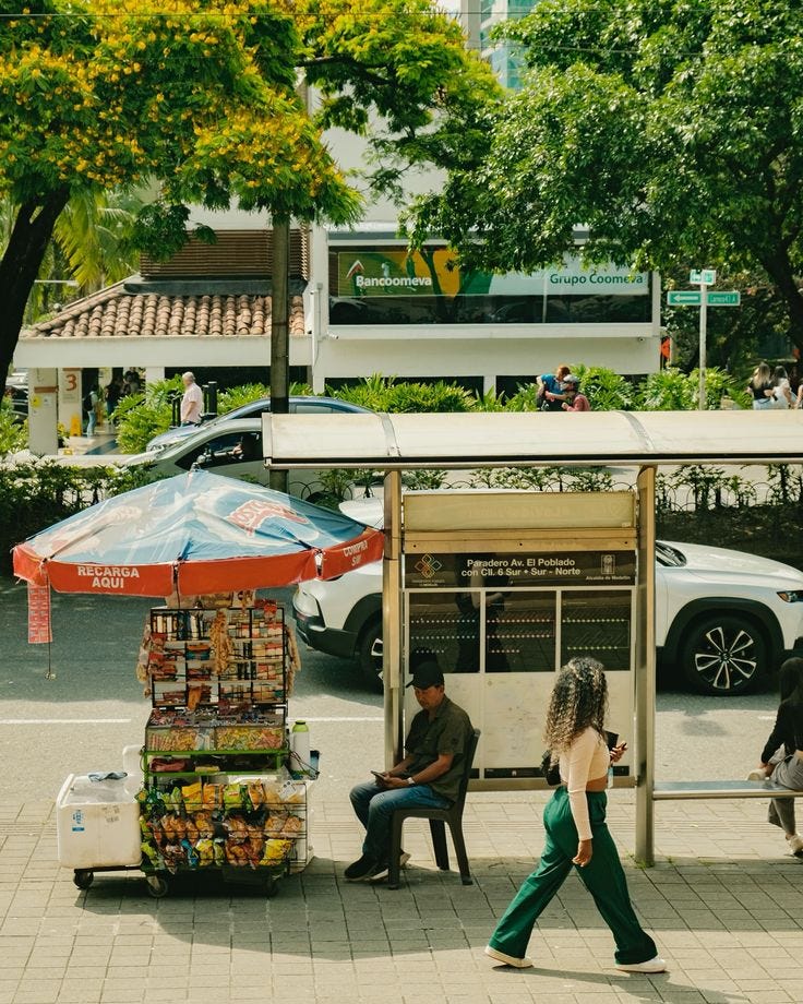 This may contain: a woman walking past a kiosk on the side of a road with people sitting under it