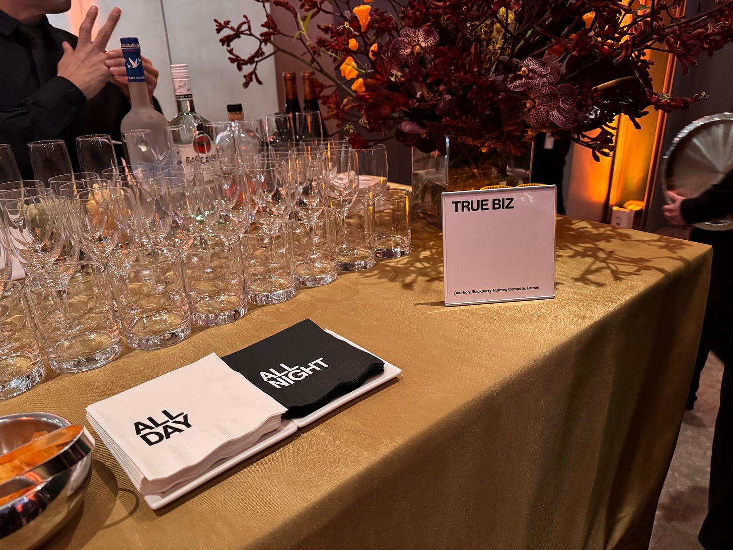 Bar glasses ready to be filled at the evening reception. Black and white napkins with "ALL DAY ALL NIGHT" are on the left-hand side, while a sign for "TRUE BIZ" drinks is on the right.