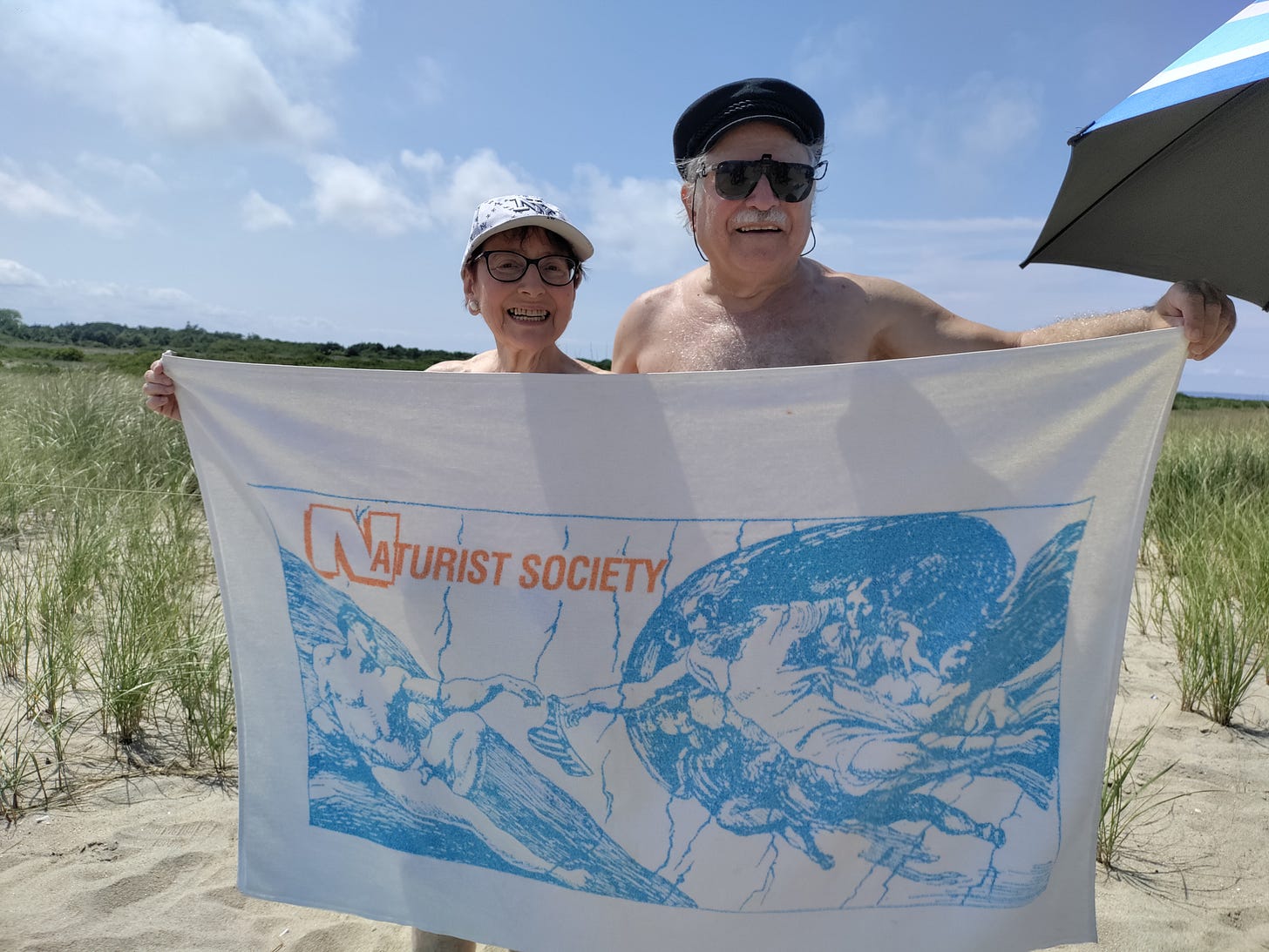 Leonard Lehrman and Helene Williams smile while holding a vintage Naturist Society towel at Gunnison Beach, surrounded by dune grass and a blue sky. Leonard Lehrman and Helene Williams smile while holding a vintage Naturist Society towel at Gunnison Beach, surrounded by dune grass and a blue sky.