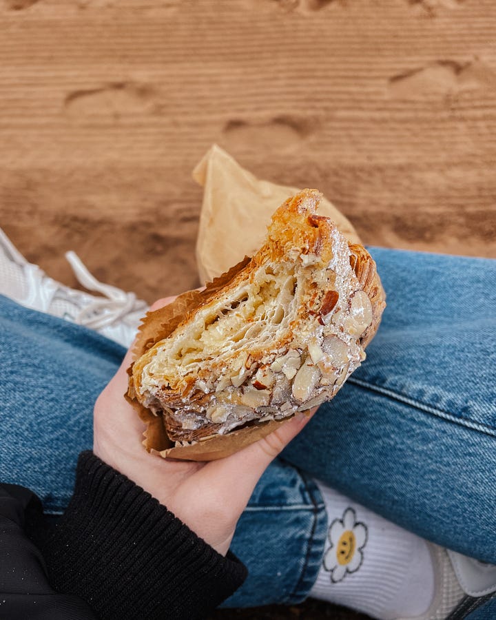 Image 1: Holding an almond croissant with a bite taken out, sitting cross-legged. Sand is visible below. / Image 2: The tide rolling in, gentle waves meeting the shore. / Image 3: Inside Tills & Company bookshop—books, tables, and people enjoying a slow moment. / Image 4: A coffee table in Tills with my book Ways of Seeing and a coffee cup resting on it.