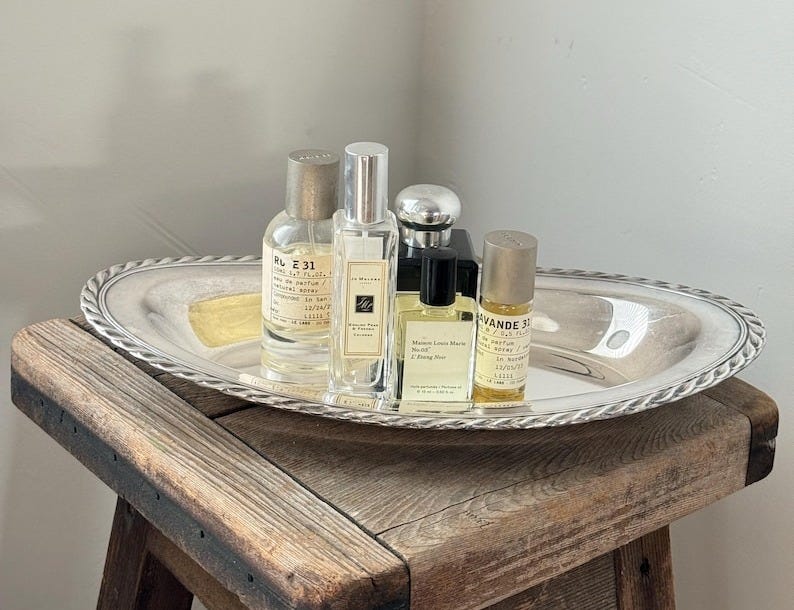 May include: A silver oval tray with a rope-like border holds several perfume bottles of varying sizes and shapes. The bottles are clear, with labels in black and white. The tray sits on a rustic wooden stool.
