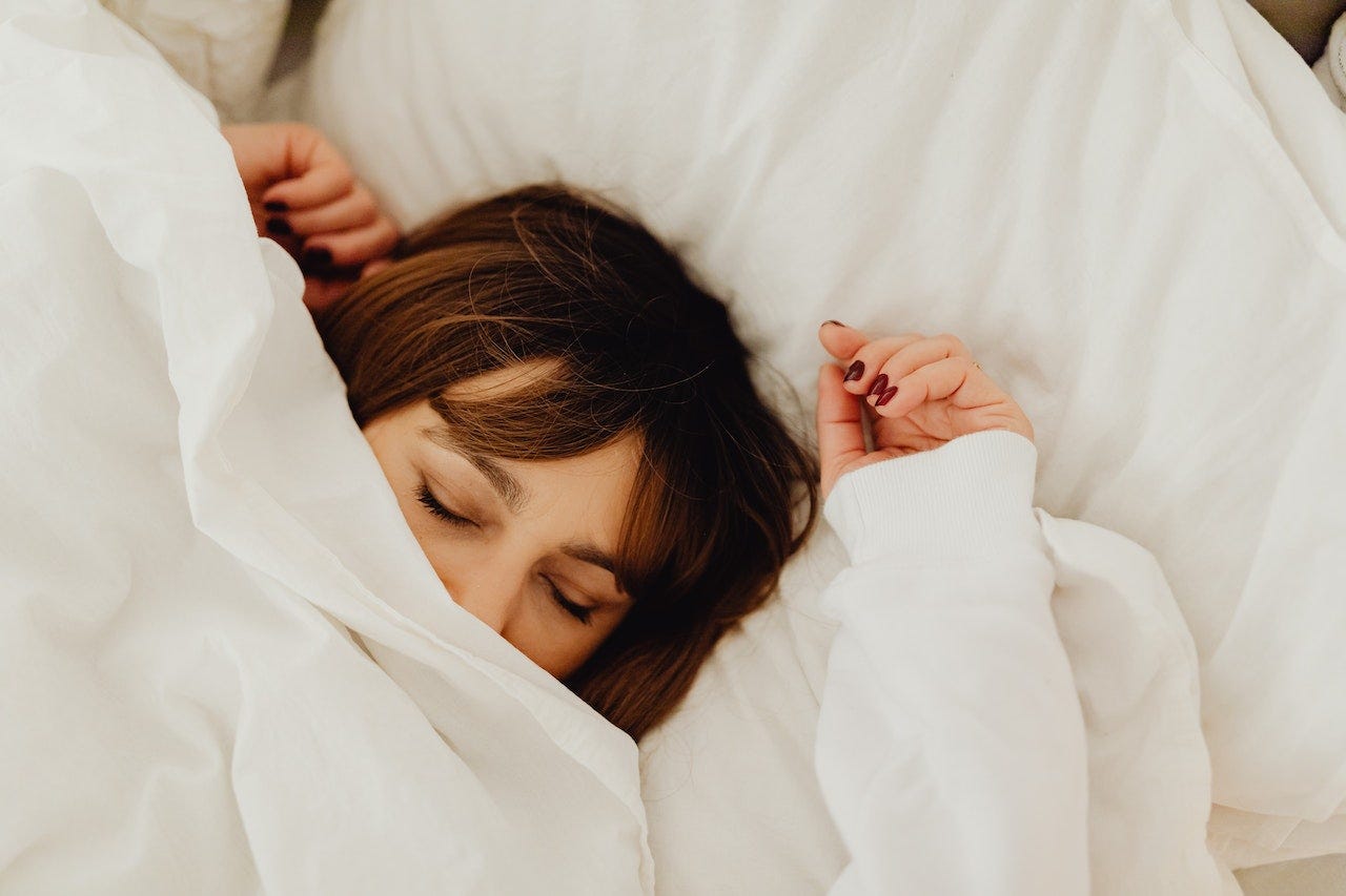 Woman in white laying in a bed made with white sheets with the top sheet pulled hallway up her face. Woman in white laying in a bed made with white sheets with the top sheet pulled hallway up her face.