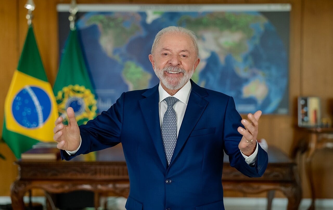 Lula stands in a dark suit with his arms open, with Brazilian flags and a world map behind him.