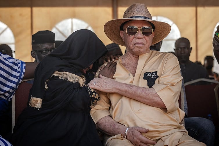 A woman in black mourning dress hides her face on the shoulder of an albino man in sunglasses and a hat, who comforts her. A woman in black mourning dress hides her face on the shoulder of an albino man in sunglasses and a hat, who comforts her.