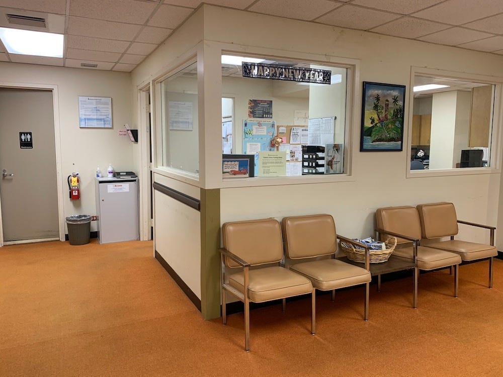 Waiting room which features a booth where the receptionist will sit, in front of which are for knackered-looking chairs in leather with a table in between them and a basket of magazines. There is also a toilet and a waste paper basket off to the left and a fire extinguisher. There is a painting on the wall and a Happy New Year banner up in one of the kiosk windows. It is clean and tidy but very grim.