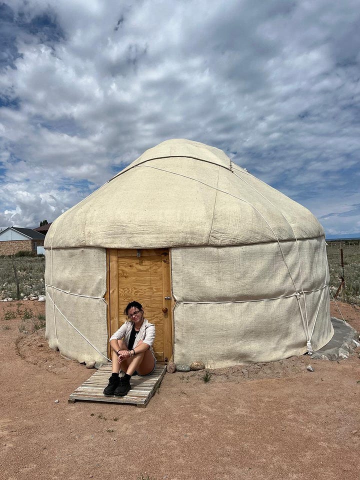 Split image: left, a woman with dark wavy hair resting her chin on her hand at a beachside café with the ocean and coastal buildings behind her; right, the same woman sitting on the wooden step of a white yurt under a partly cloudy sky.