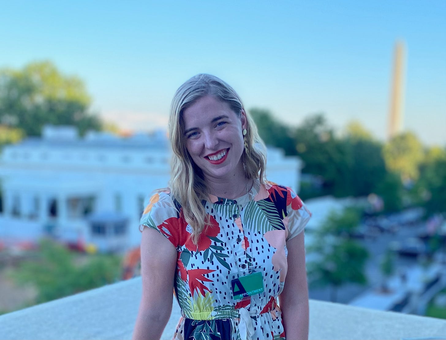 Catie, a blonde woman in her 30s with red lipstick standing on a balcony facing the camera. Behind her is the White House and Washington monument.