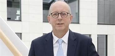 Un hombre con traje y gafas posa frente a un edificio moderno.
