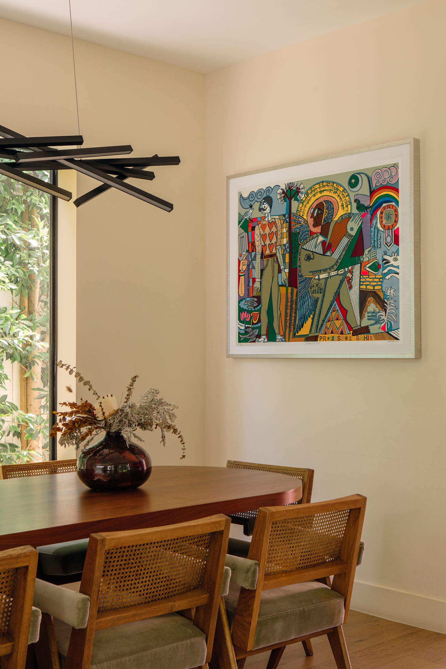 Dining room with a walnut oval table, cane-back chairs and a black linear chandelier. Jyoti Bhatt's Myself As An Artist, a large, colorful serigraph with a wide white mat and pale frame, hangs on the cream wall.