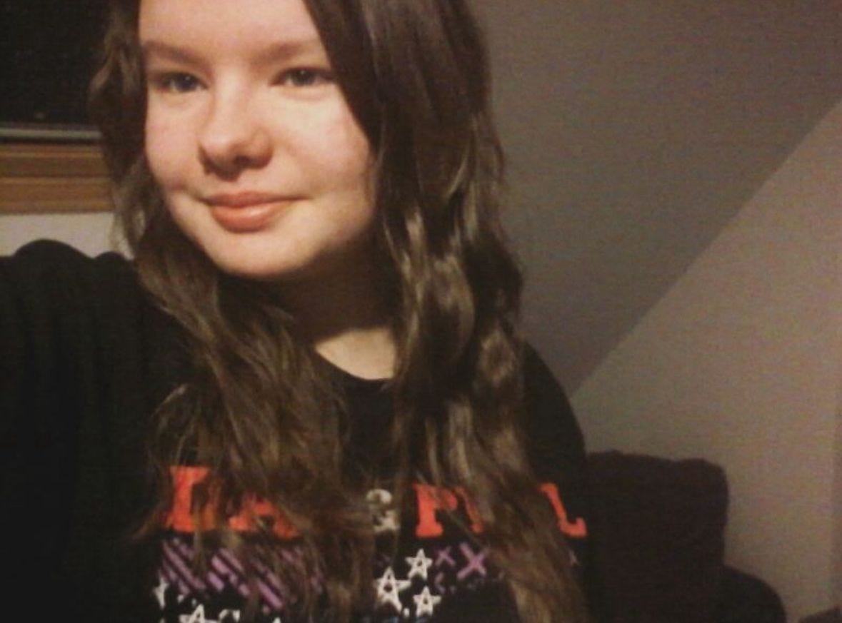 Charli in 2016 - a white teenager with brown hair wearing a Dan and Phil tour tshirt that peeks behind their hair. They are smiling but it doesn't reach their eyes. The photo is quite filtered making it quite sepia toned