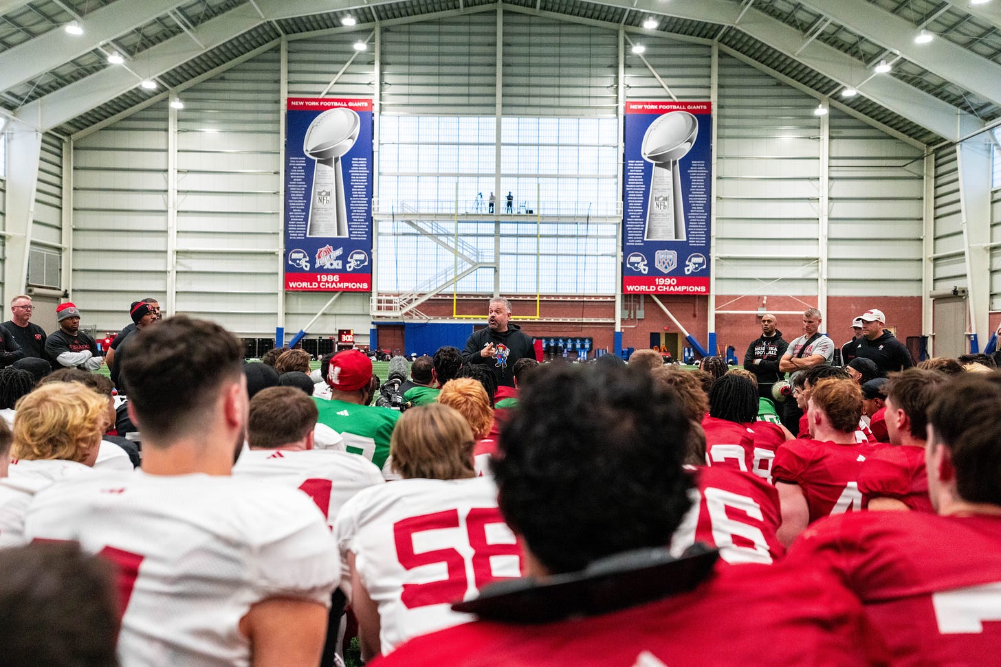 Coach Matt Rhule addressing a group of players wearing red and white jerseys during an indoor practice session, with banners celebrating the New York Giants' championships prominently displayed in the background.