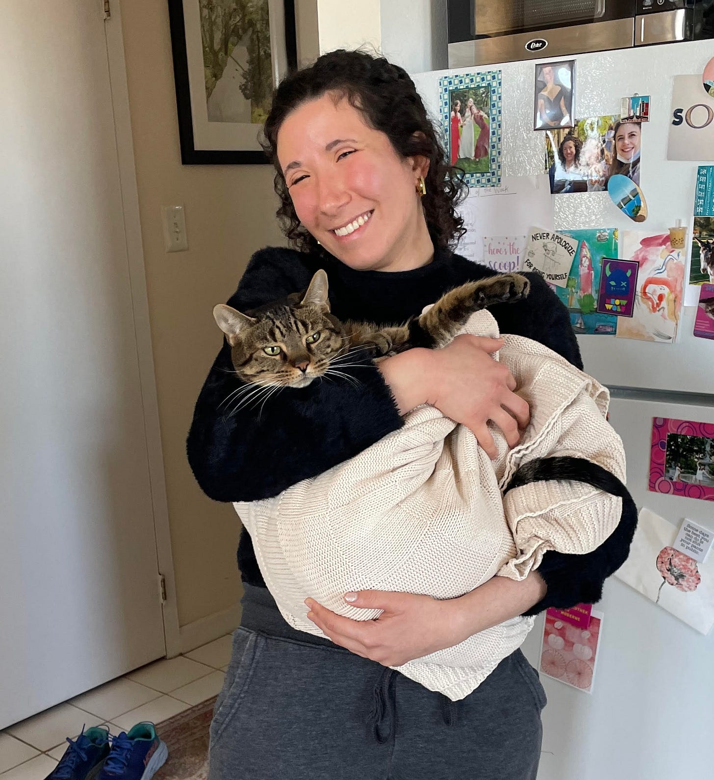 A woman standing in front of a fridge, smiling and holding a cat wrapped in a blanket. The cat looks relaxed and comfy in the blanket. The girl is wearing a black sweater and gray sweatpants. She has curly black hair in a ponytail.
