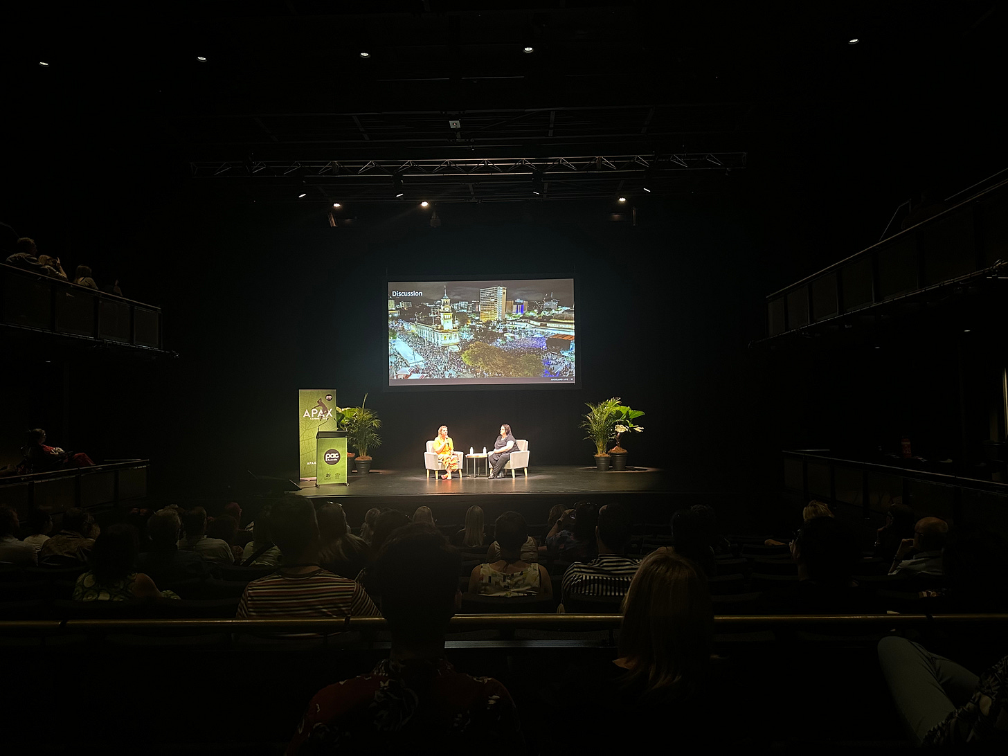 A wide shot of a conference stage with two speakers seated in armchairs, engaged in discussion. A large screen behind them displays an image, while the audience is seated in a dark auditorium. The room is dimly lit, except for the bright stage lights, and potted plants are positioned on either side of the stage for decoration.