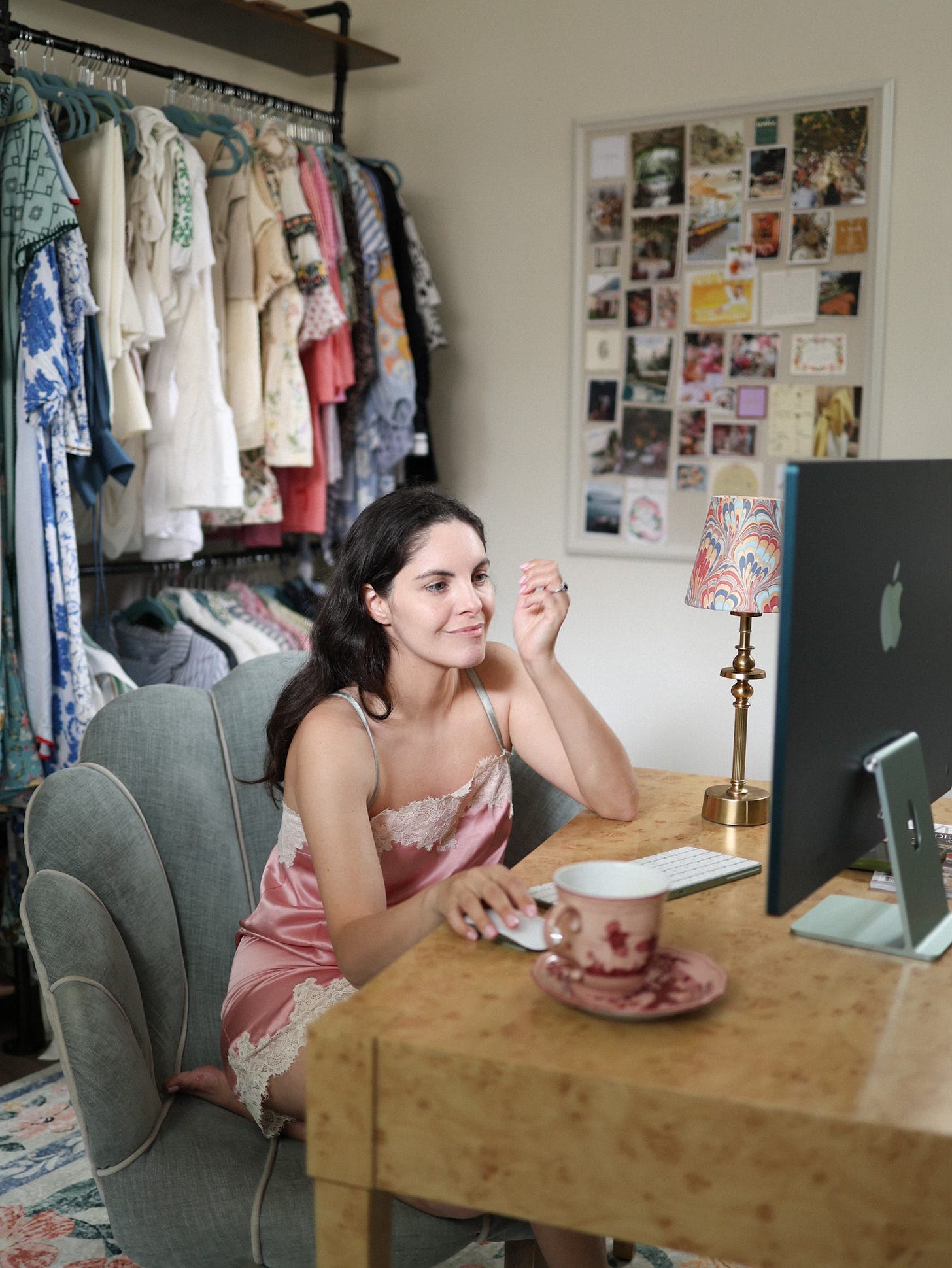 Monica at her wooden desk in her closet office, morning light, holding coffee mug, with clothing rack and mood board visible in background, during her 5am writing practice.