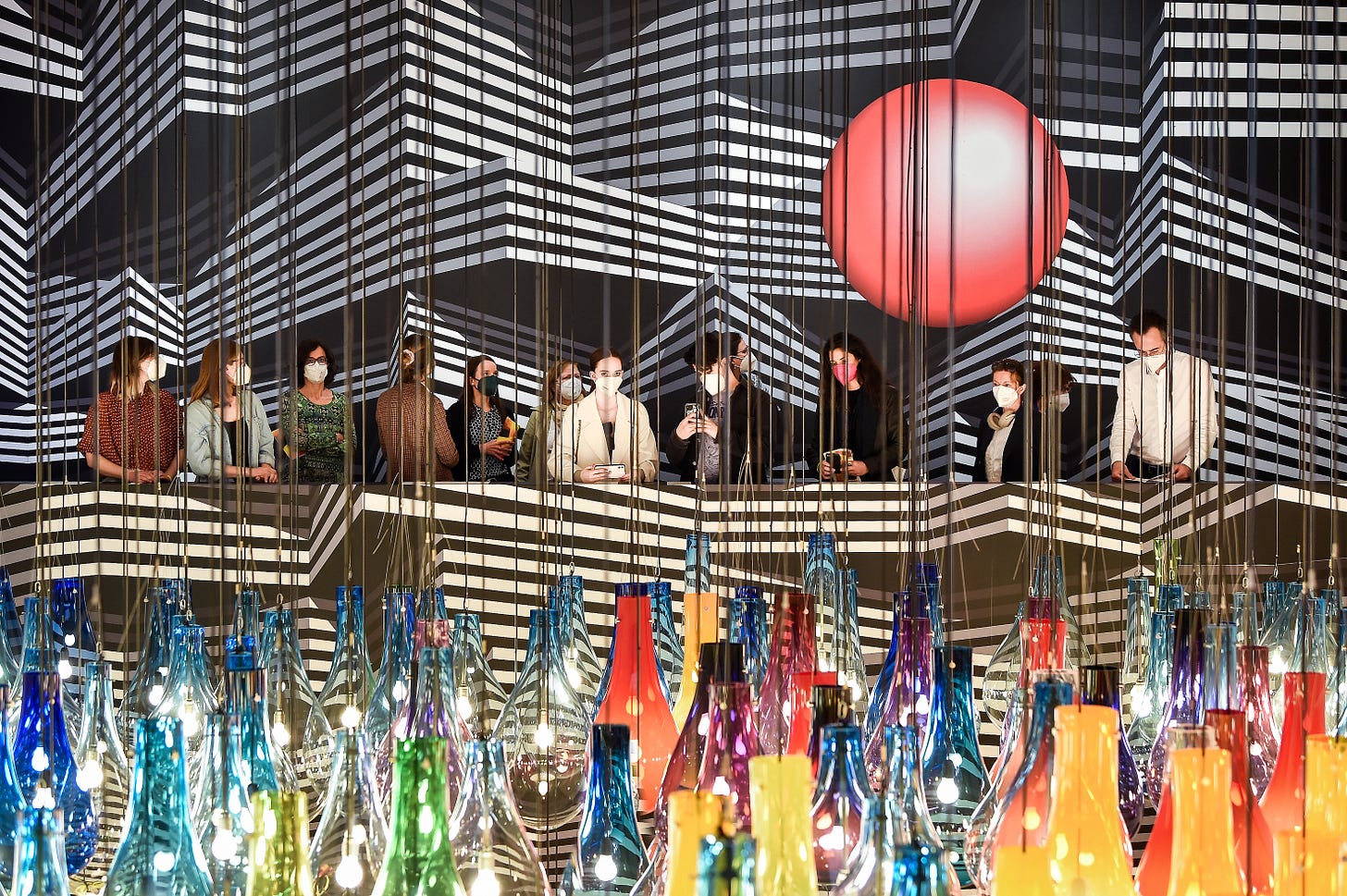 Masked visitors at the Tobias Rehberger exhibition. They are standing in a gallery painted with black and white stripes; in front of them is hanging lots of large glass lightbulbs in different colours Masked visitors at the Tobias Rehberger exhibition. They are standing in a gallery painted with black and white stripes; in front of them is hanging lots of large glass lightbulbs in different colours