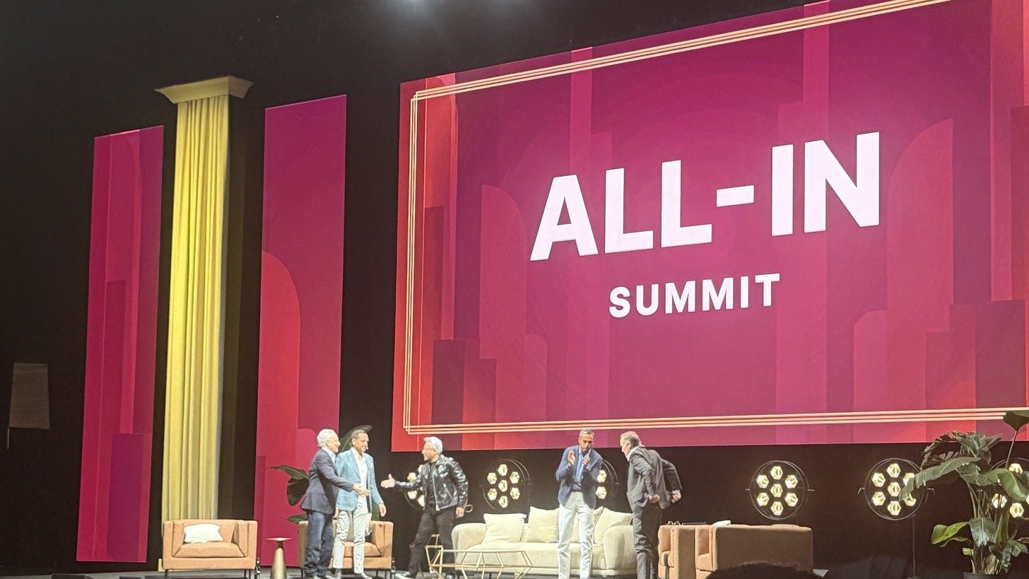 The All-In Summit stage at The Shrine in Los Angeles, photographed from the audience: a large red screen reading ALL-IN SUMMIT above a lounge set where the four hosts, in jackets, greet Jensen Huang in his black leather jacket at center.