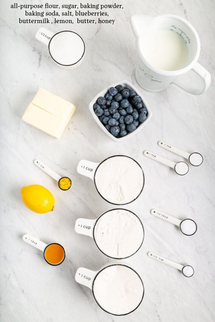 Overhead view of baking ingredients on a white surface: flour, sugar, baking powder, baking soda, salt, buttermilk, blueberries, butter, honey, a lemon, and measuring spoons and cups filled with ingredients. Overhead view of baking ingredients on a white surface: flour, sugar, baking powder, baking soda, salt, buttermilk, blueberries, butter, honey, a lemon, and measuring spoons and cups filled with ingredients.