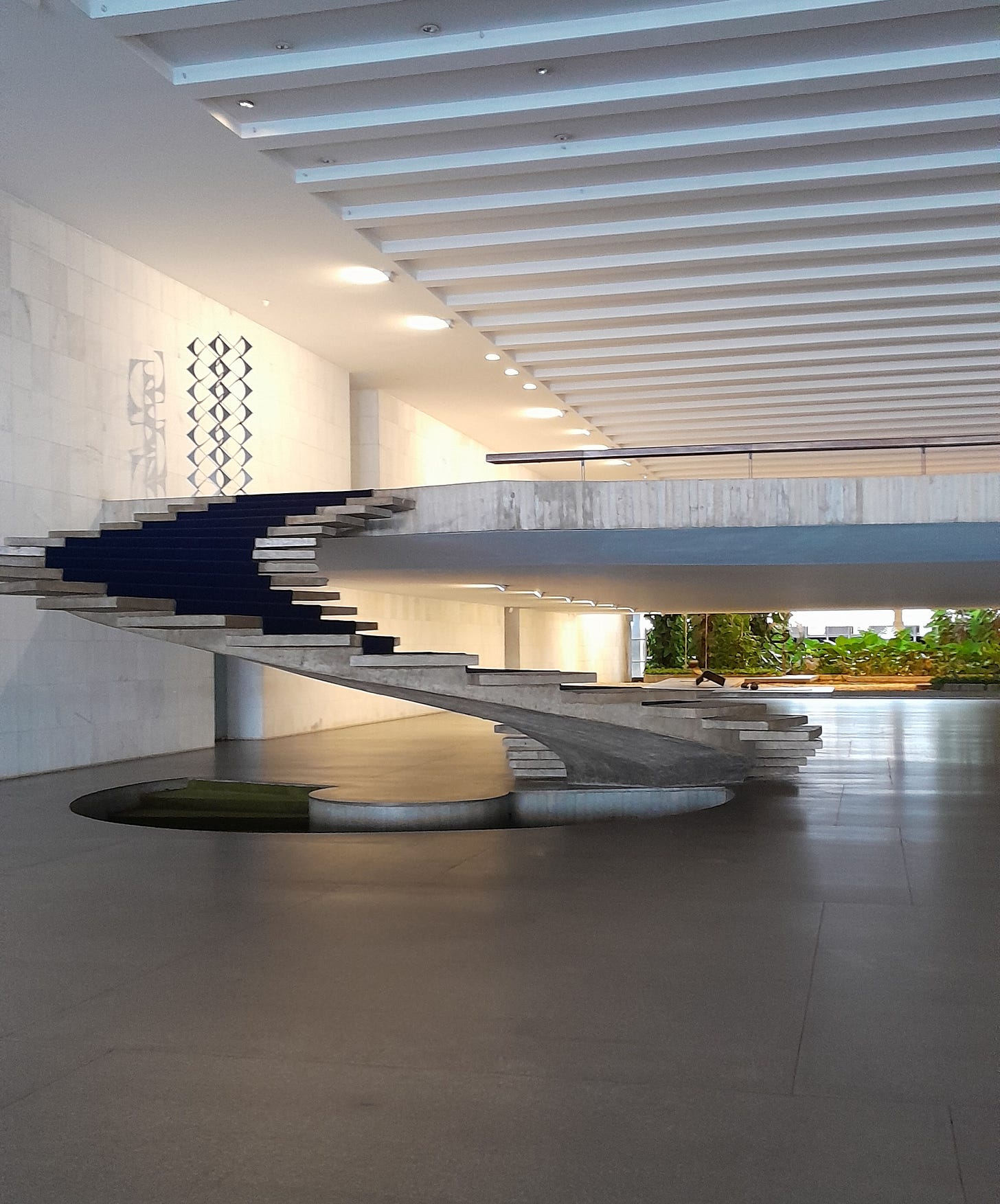 Um vão comprido com uma escadaria curva e larga, sem corrimão. Ela faz uma curva de 90º para levar ao piso de cima, no qual podemos ver uma espécie de escultura. Ao fundo, algum jardim.