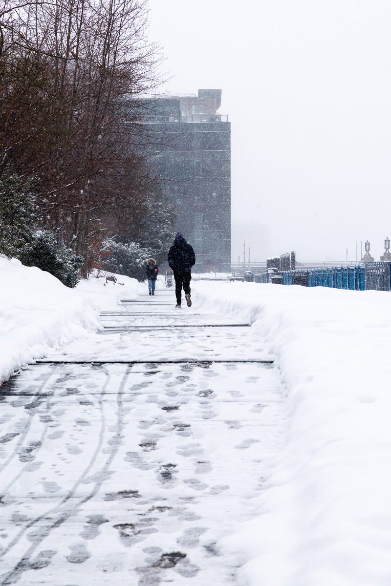 Pedestrians walking along a snow-covered HarborWalk with fresh footprints in Boston Seaport.