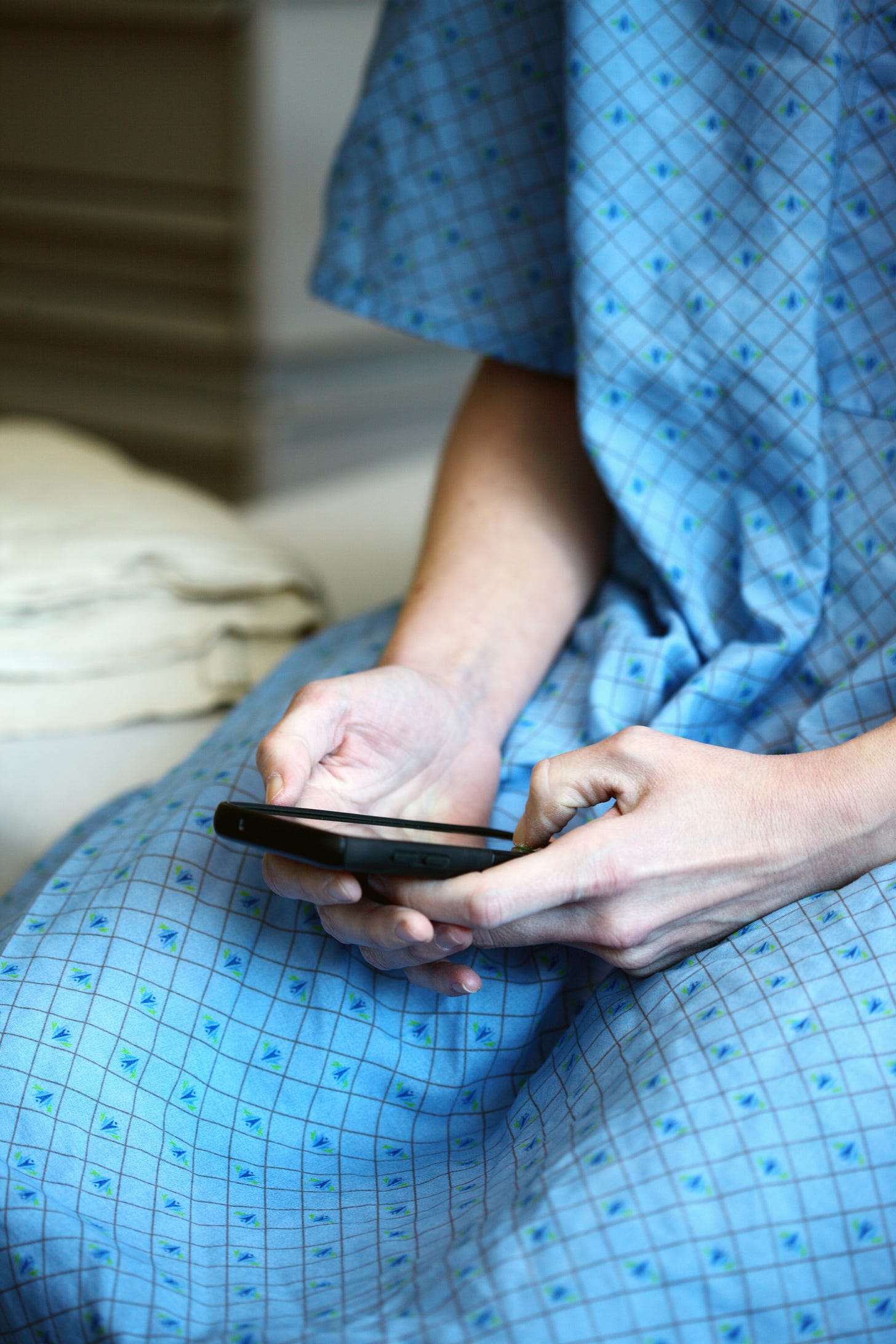 A person in a hospital gown sitting on a bed reading their cell phone. A person in a hospital gown sitting on a bed reading their cell phone.