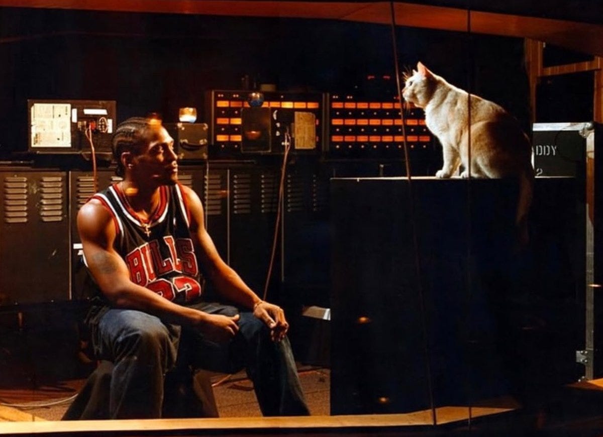 Black and white image shows D’Angelo, a Black man with dreadlocks, sitting on a stool in a recording studio. He wears a sleeveless Chicago Bulls jersey numbered 23 and baggy jeans, arms crossed relaxed. A beige cat sits on top of audio equipment to his right. The studio features control panels with knobs and screens, hanging cables, metal lockers, and a wooden floor. Dim lighting from overhead lamps creates shadows.